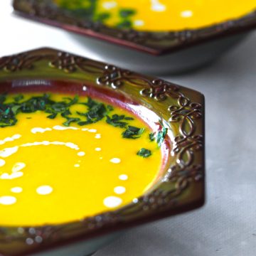 a brown bowl filled with yellow soup topped with green herbs and white drops