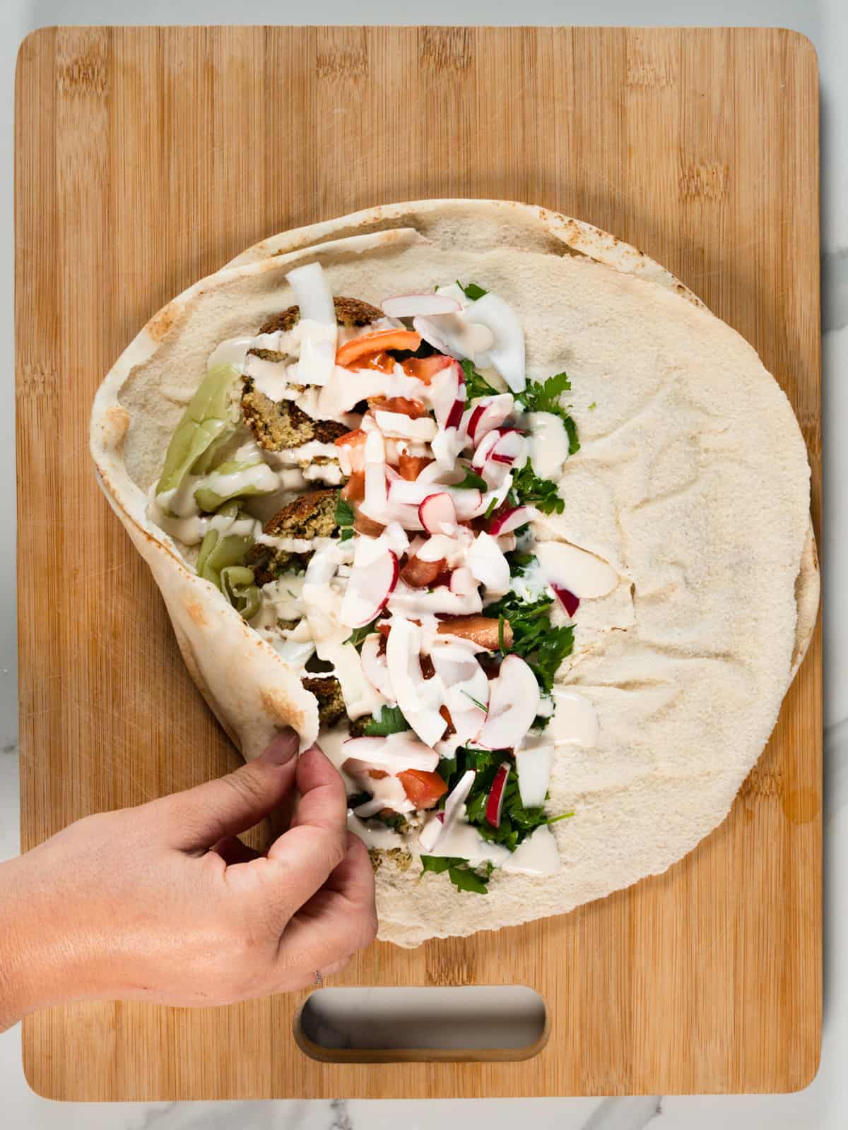 a female hand holding the edge of a round pita bread filled with chopped veggies, white sauce and squashed falafel