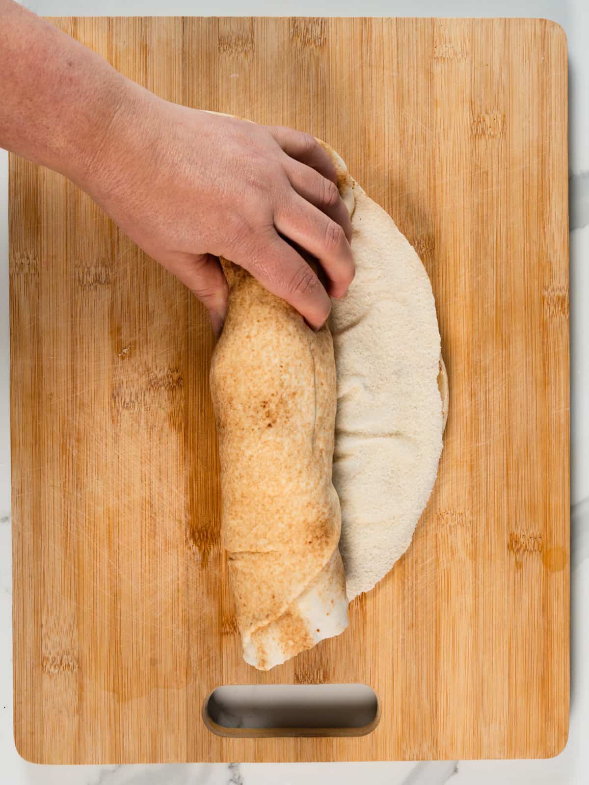 a female hand rolling a pita bread sandwich roll on a wood chopping board