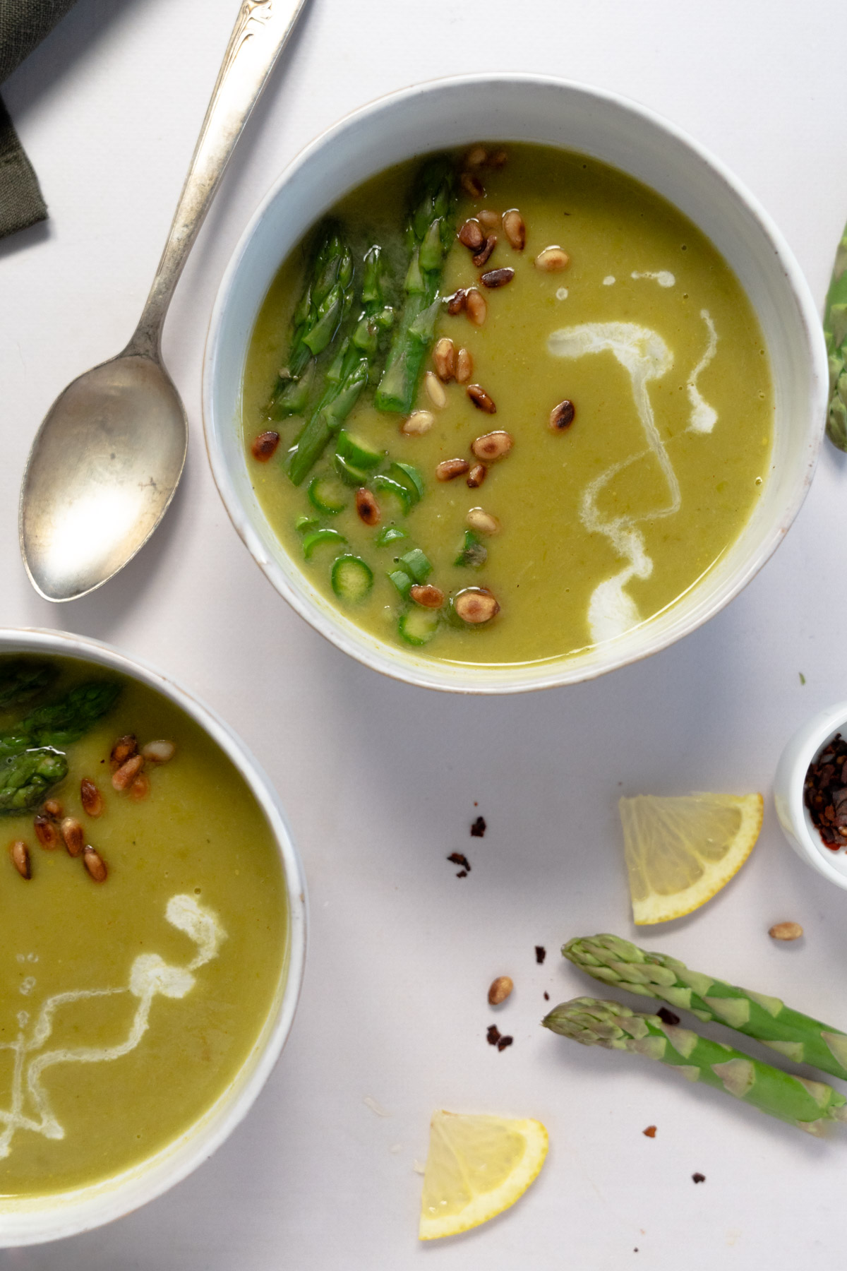two white bowls of light green soup topped with pine nuts, a drizzle of white coconut milk and chopped 
asparagus