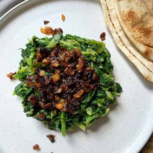 a white plate with steamed greens topped with fried onion and a side of Lebanese bread