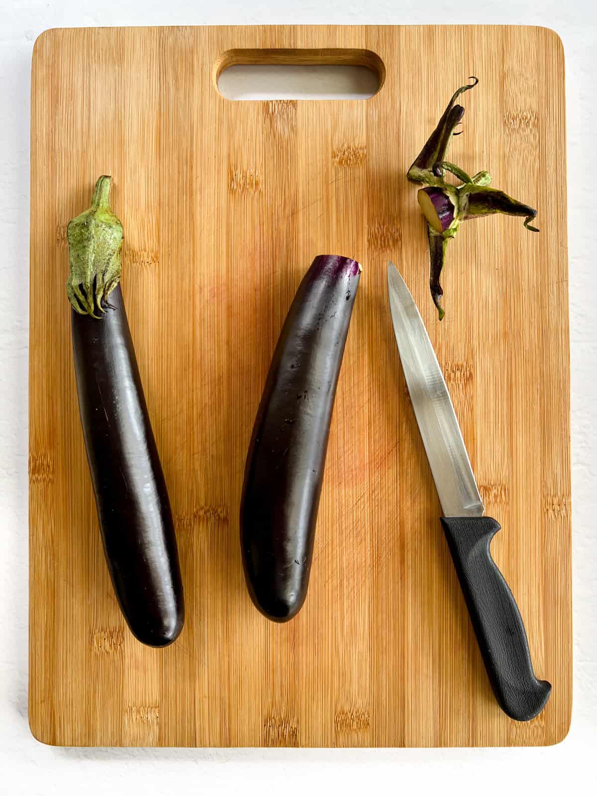 two long eggplants one with its top cut off and a knife on a timber chopping board