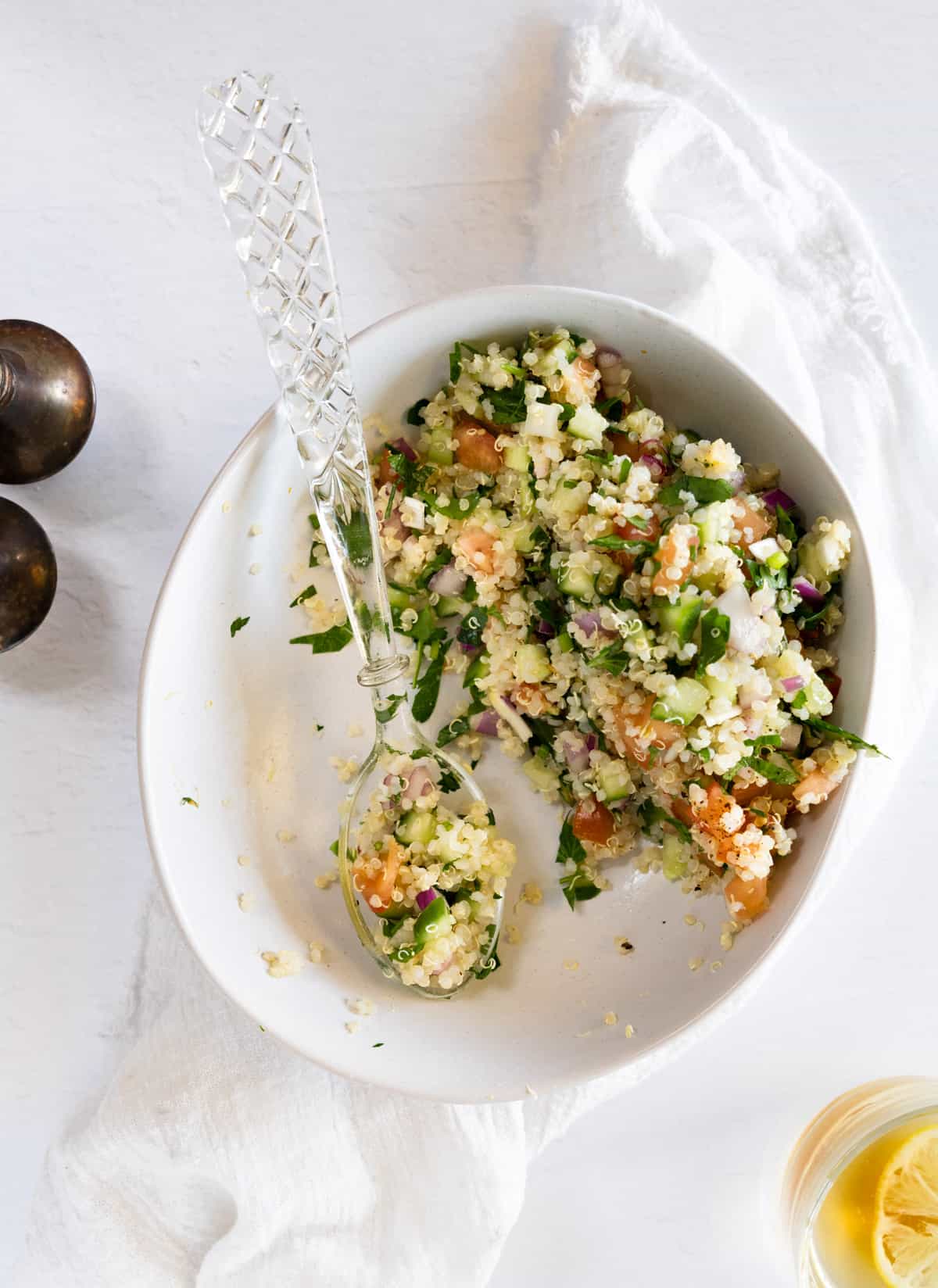 A white bowl with a glass spoon filled with quinoa and chopped vegetables