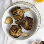three roasted artichokes in a white plate with a side of lemon