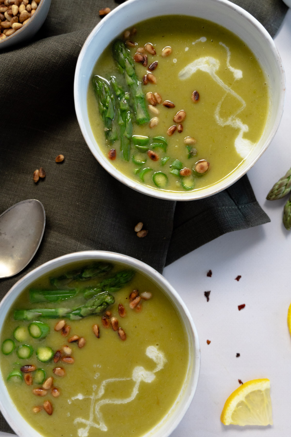 two white bowls of green asparagus soup topped with chopped asparagus and toasted pine nuts