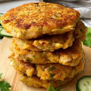 A stack of 5 sweet potato & zucchini fritters on a chopping board with cucumber slices on the sides
