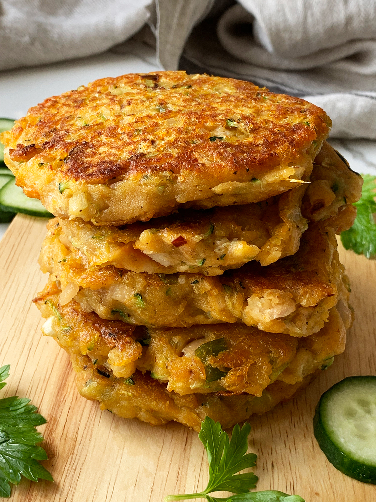 A stack of 5 sweet potato & zucchini fritters on a chopping board with cucumber slices on the sides