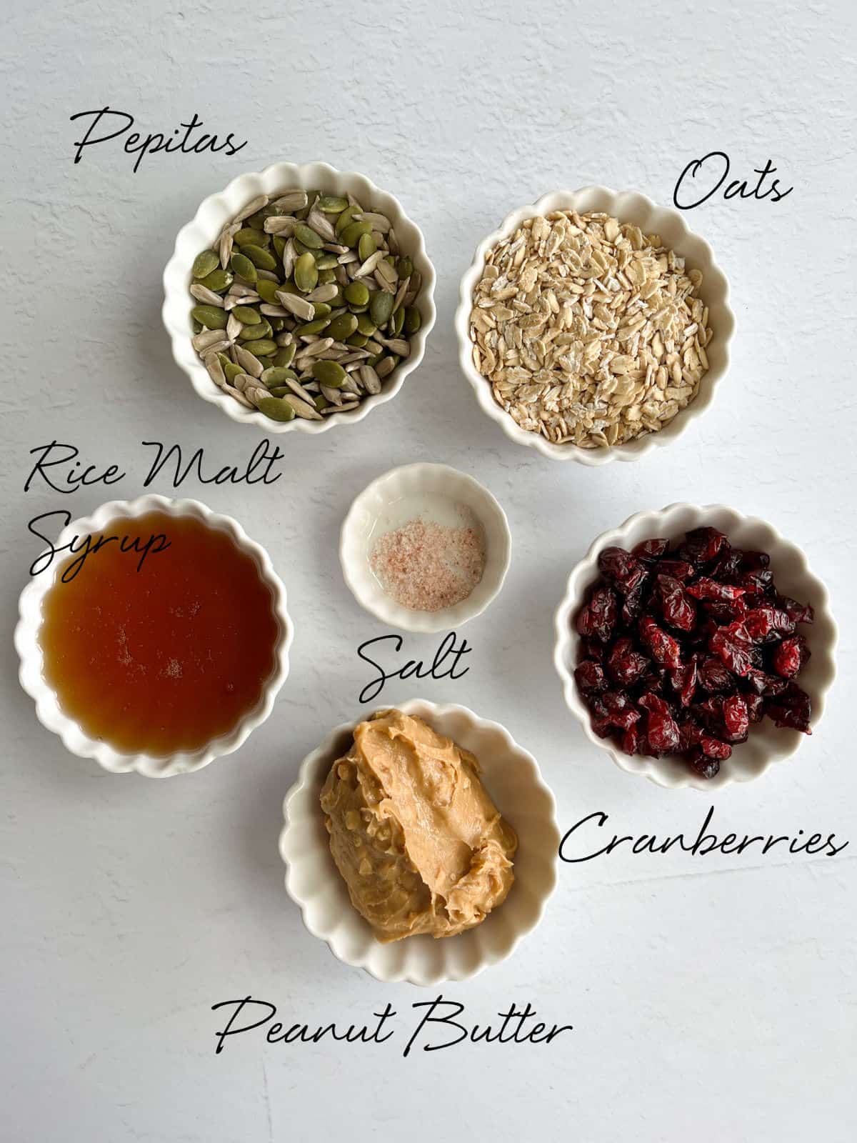 seeds, oats, a brown syrup, cranberries, salt and peanut butter in white bowls
