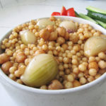 Lebanese Pearl Cous Cous in a white bowl with a side of cucumber and tomato in the background