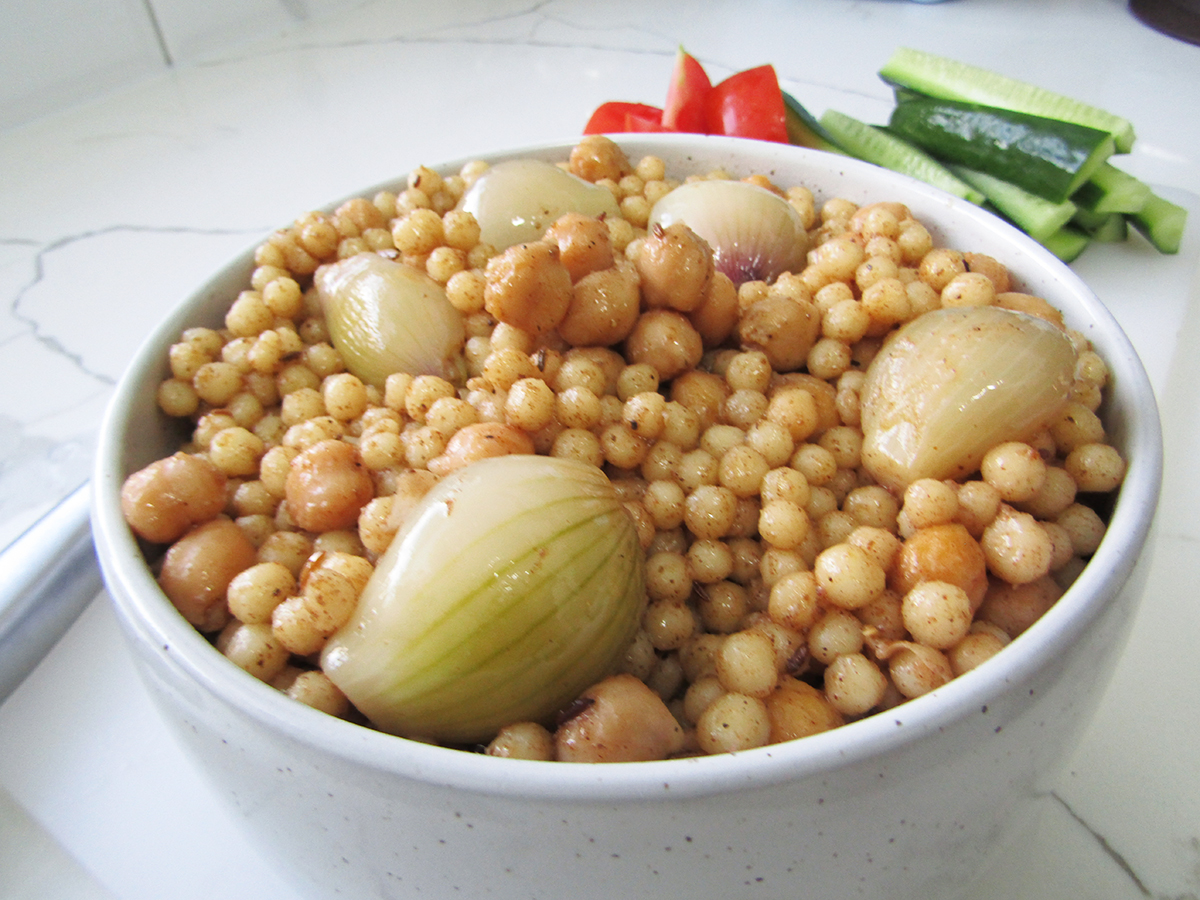 Lebanese Pearl Cous Cous in a white bowl with a side of cucumber and tomato in the background