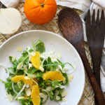 Orange and Fennel Salad (vegan recipe) served on a white plate with an orange, half onion and salad serving utensils in background