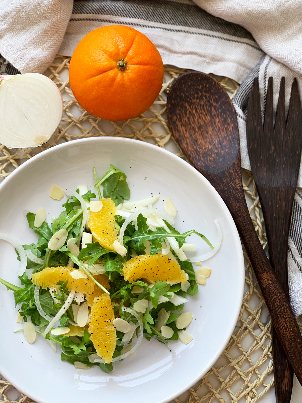 Orange and Fennel Salad (vegan recipe) served on a white plate with an orange, half onion and salad serving utensils in background