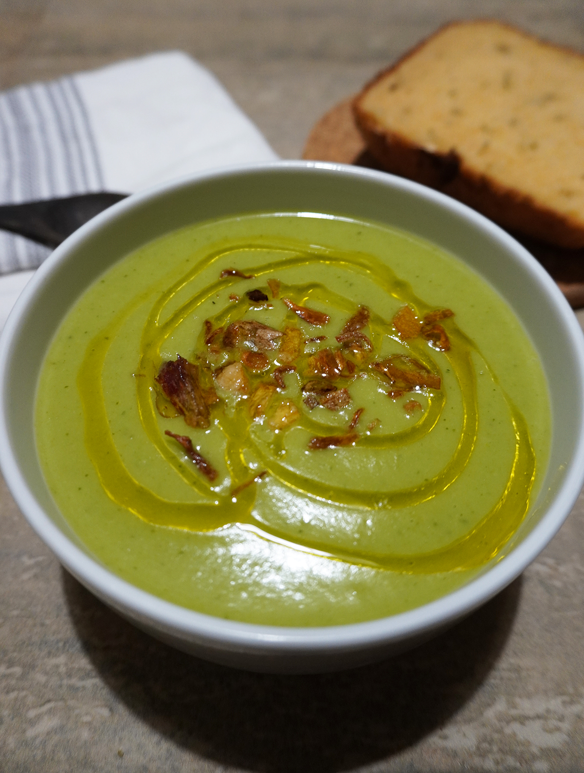 Vegan broccoli soup garnished with fried shallot and a drizzling of olive oil. Bread and spoon in the background