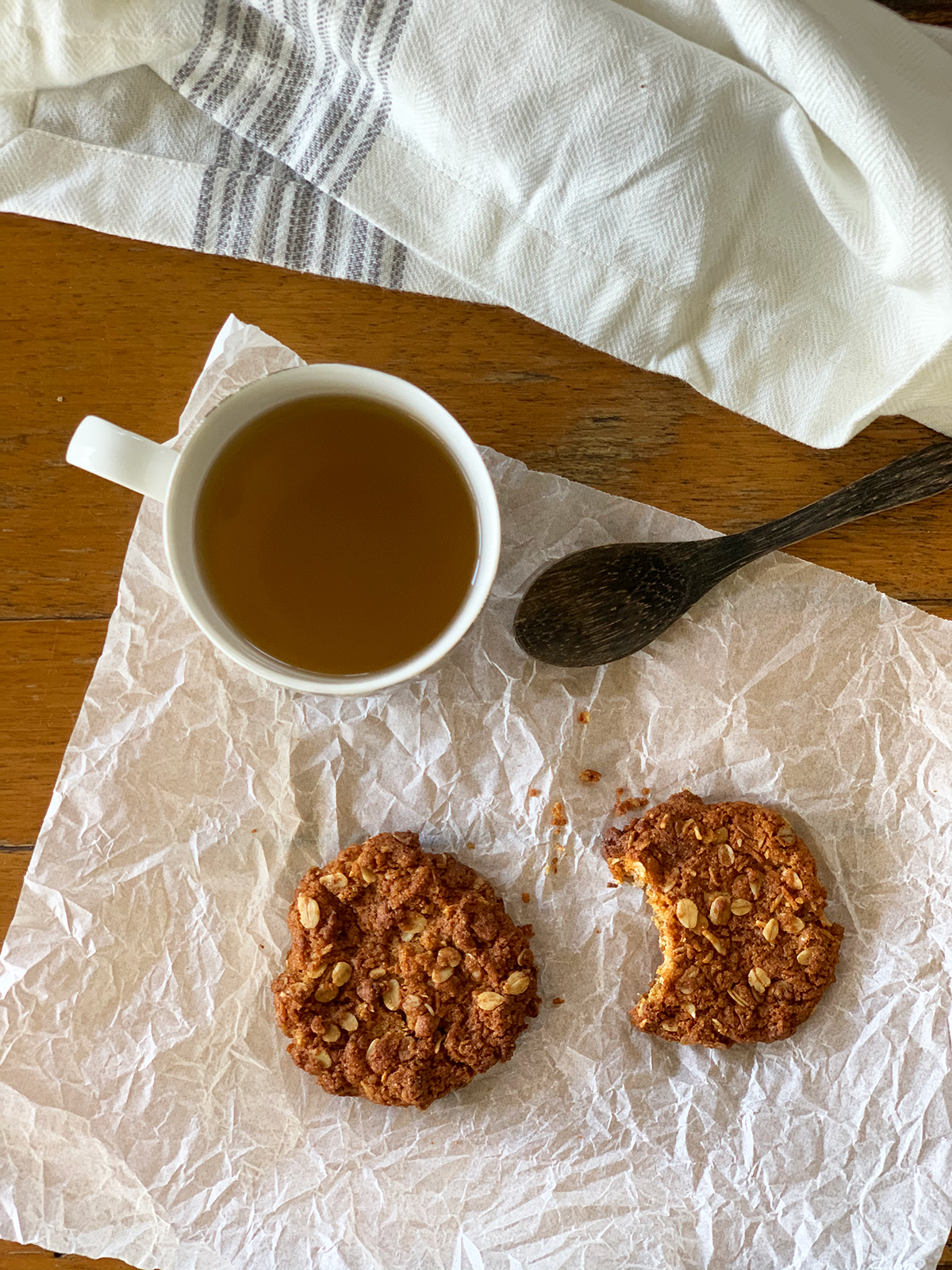Two ANZAC biscuits one with a bite out of it with a cup of ginger lemongrass tea