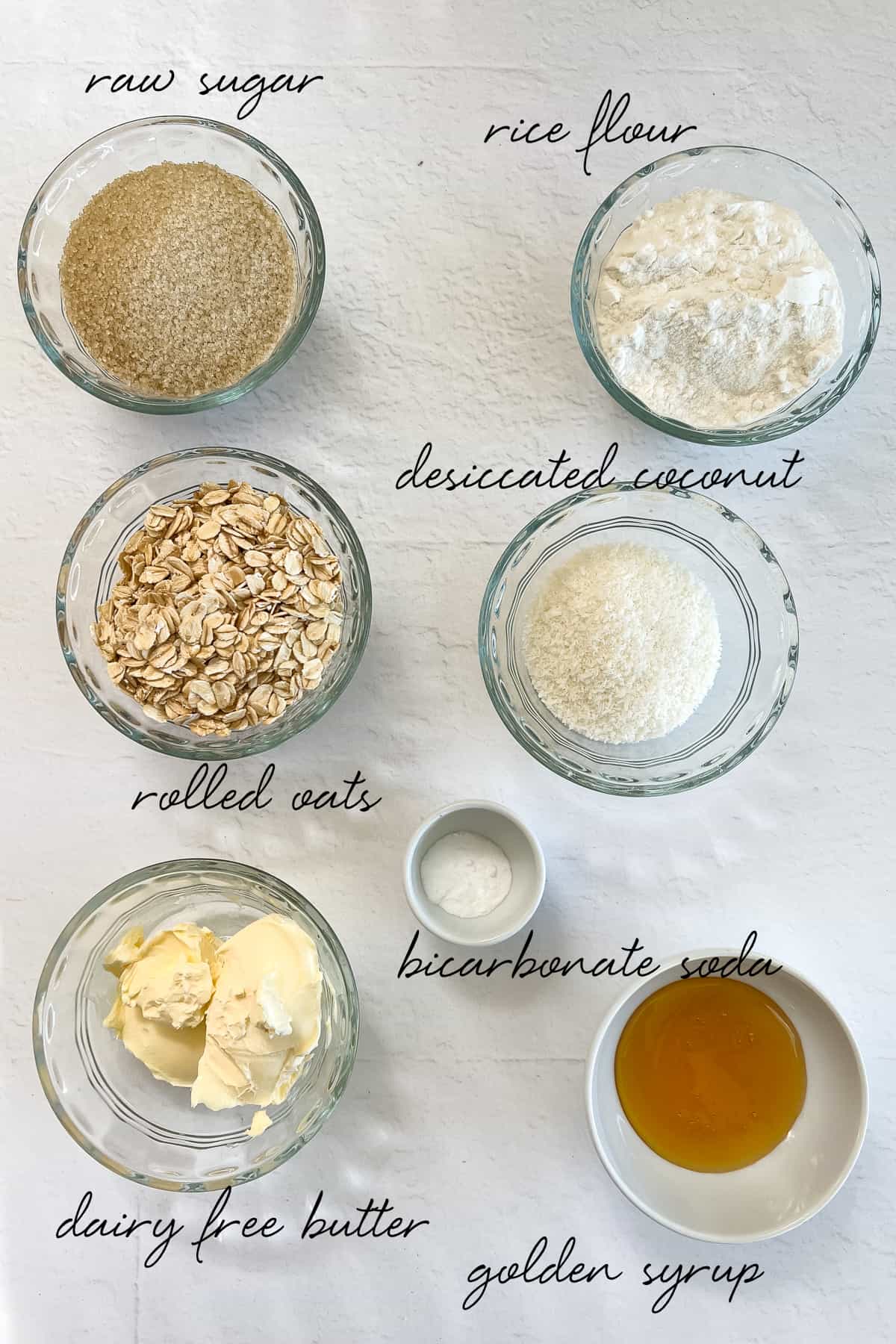 raw sugar, rice flour, rolled oats, desiccated coconut, dairy free butter, bicarb soda and golden syrup laid out in glass bowls
