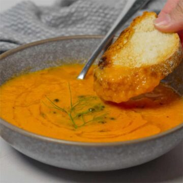 a grey bowl filled with orange soup and a piece of bread being dunked into it