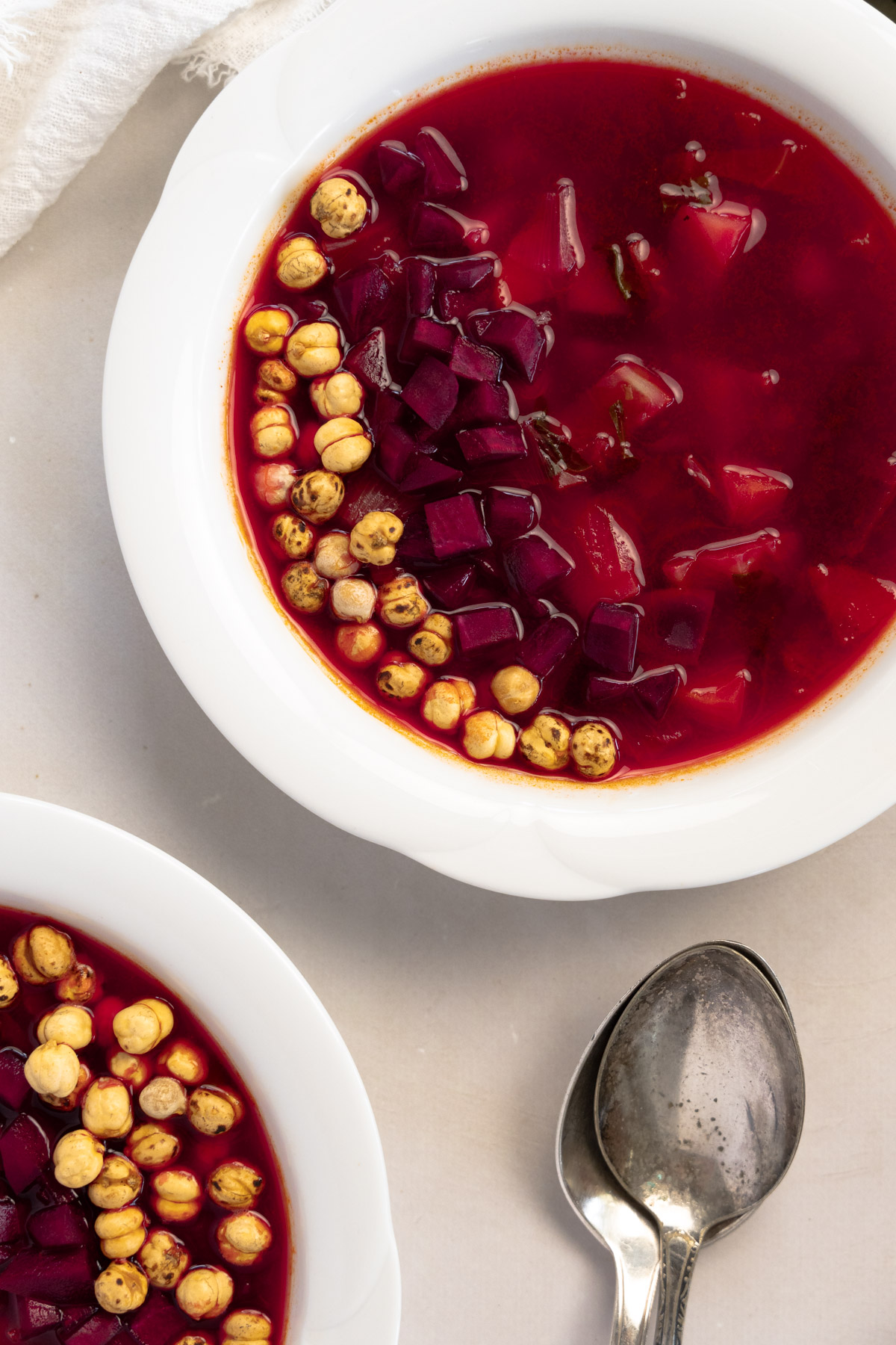 two white bowls filled with pinkish red vegetable borscht with spoons on the side