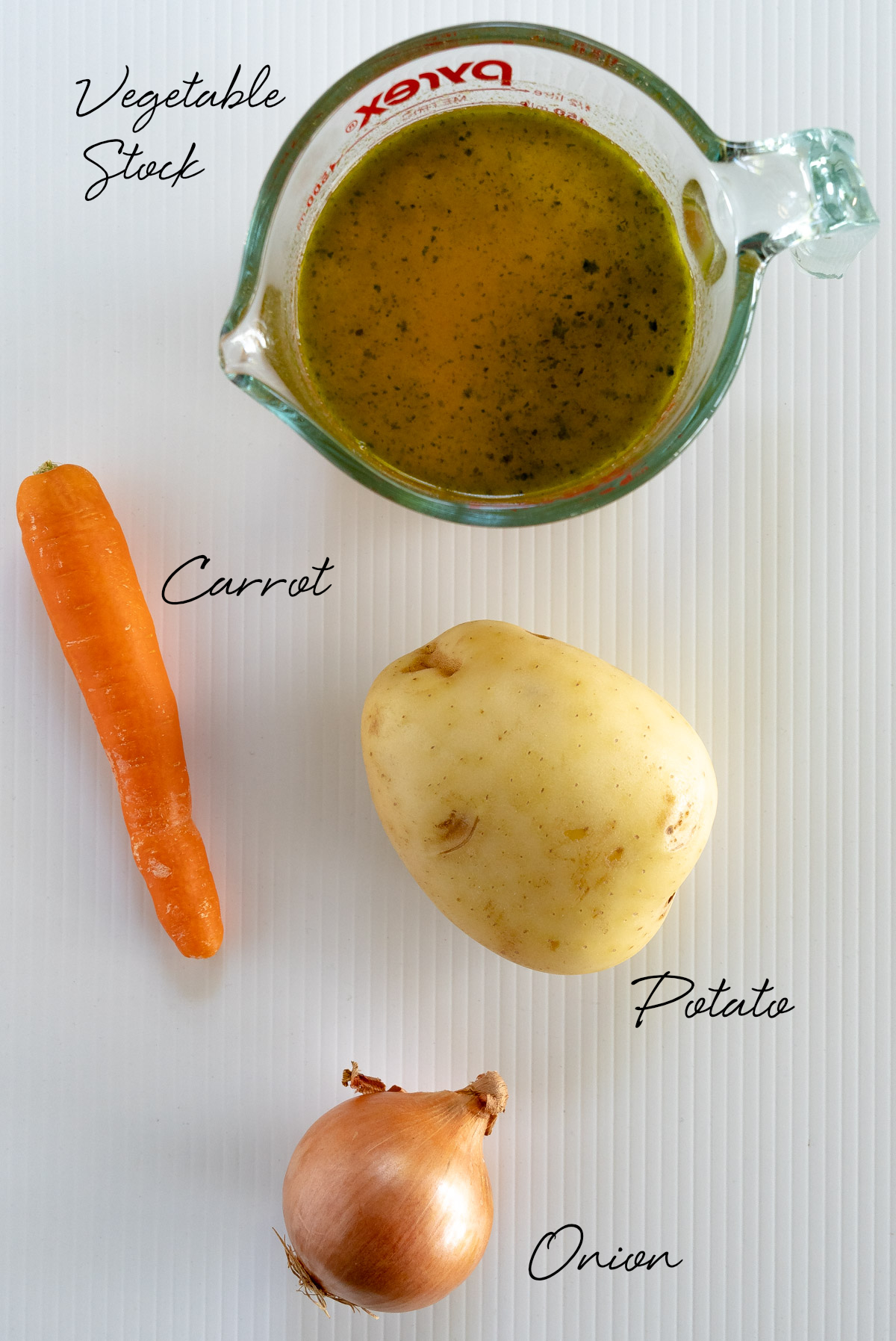 vegetable stock in a jug, a carrot, potato and onion on a white bench