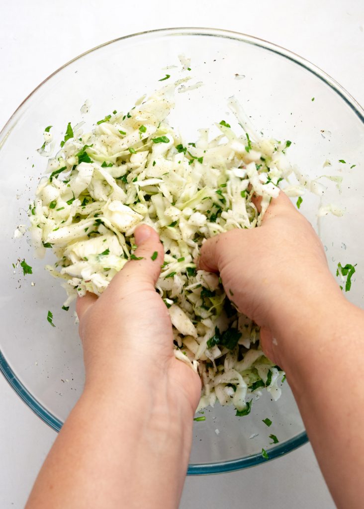 two female hands massaging shredded cabbage and parsley in a bowl