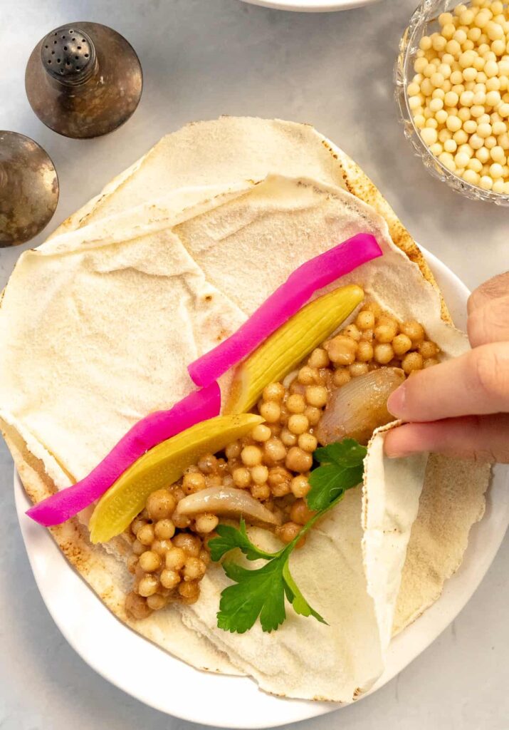 pita bread filled with mograbieh, pickles and parsley being wrapped by a male hand