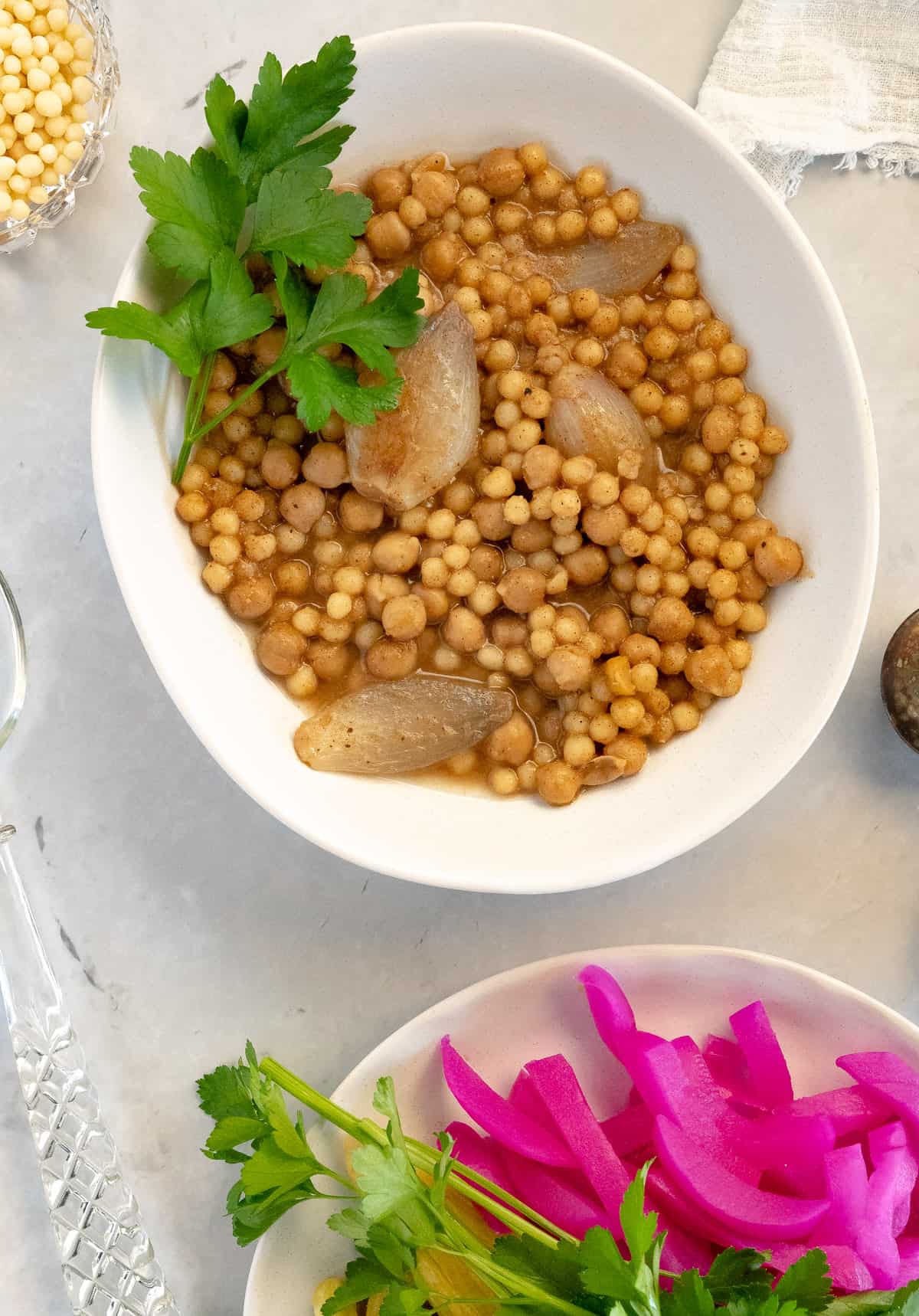 cooked pearl couscous with onions and chickpeas in a white plate with a side of pickles