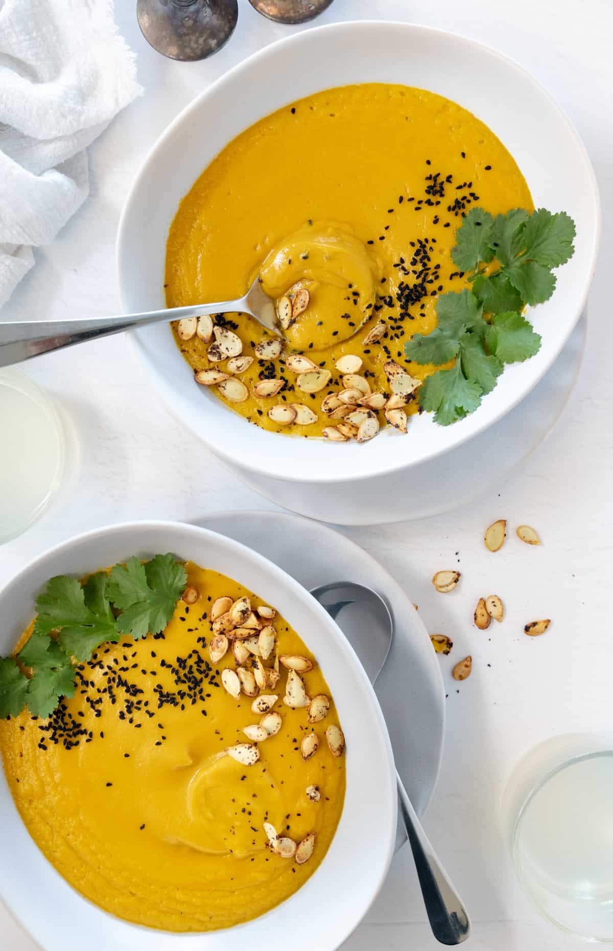 two white bowls with yellow soup topped with green herbs and toasted pumpkin seeds