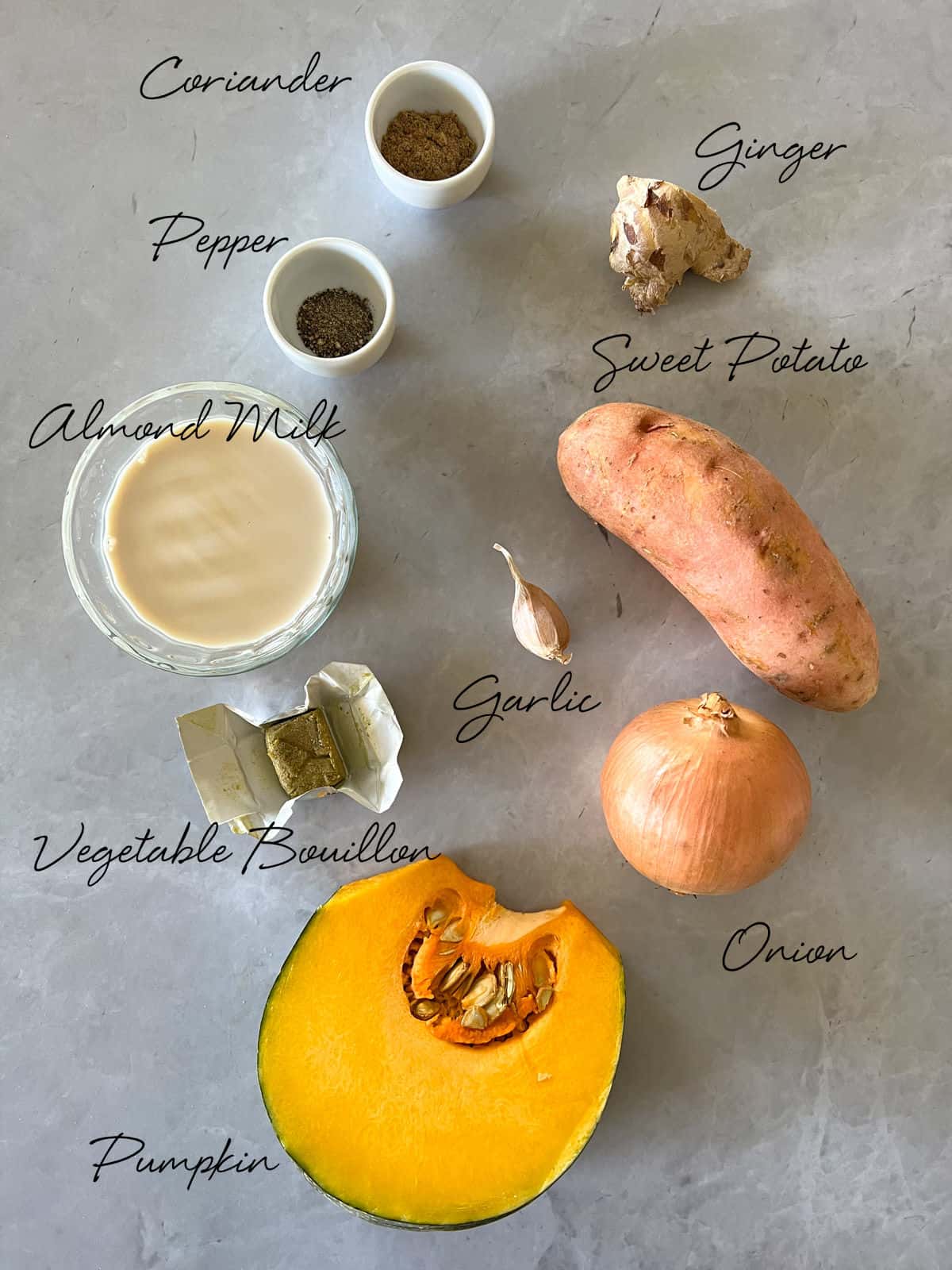a wedge of pumpkin, sweet potato, onion, garlic clove, bouillon, ginger, almond milk, pepper and ground coriander on a grey benchtop