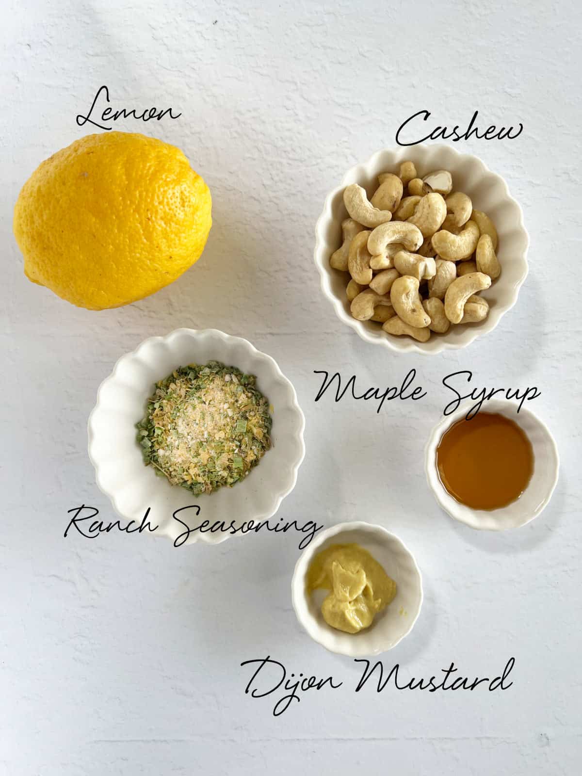 a lemon, cashews, ranch seasoning, mustard and maple syrup in bowls on a white bench top