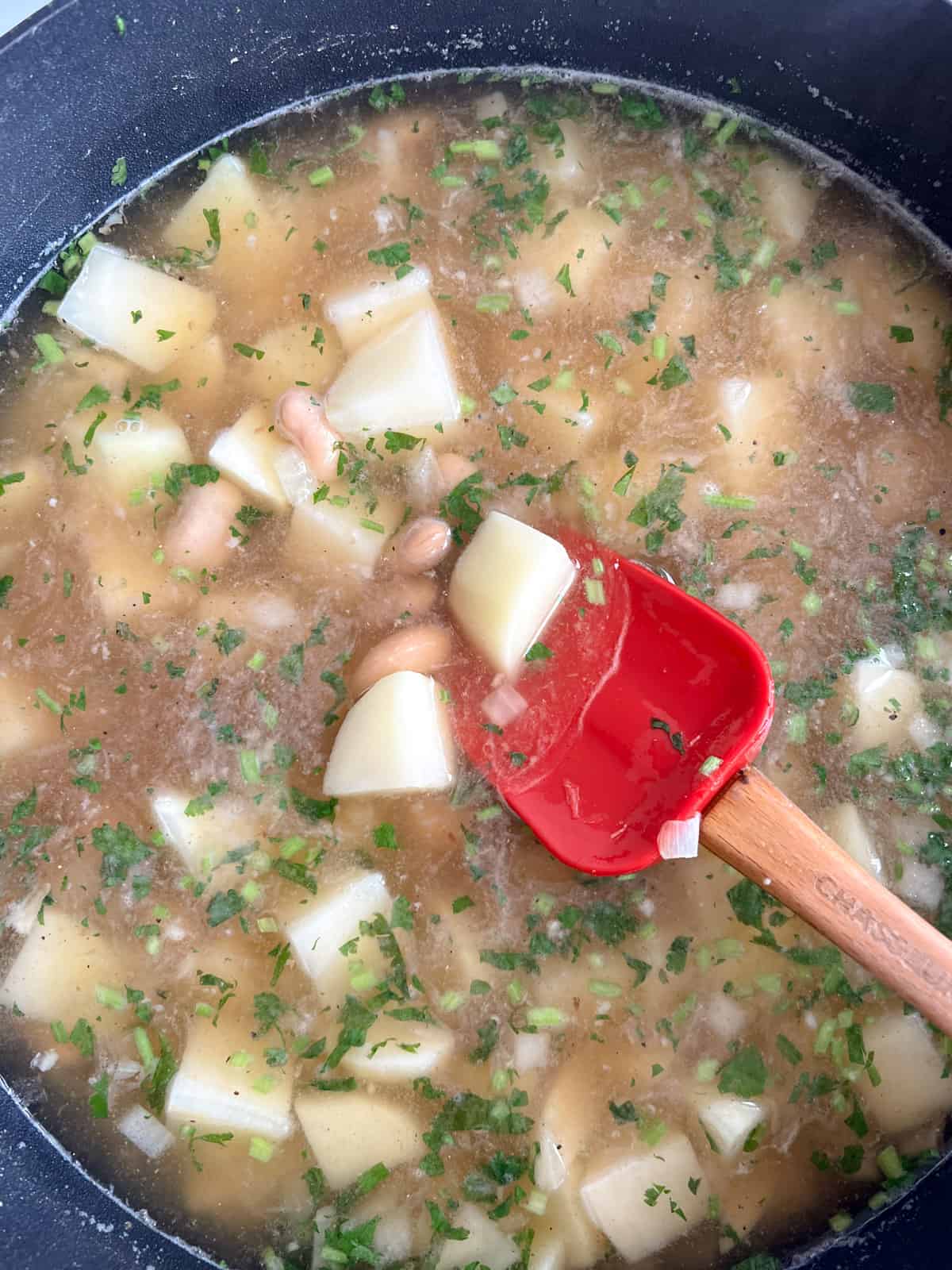 a black pot with potatoes, lima beans, herbs and onion cooking and a red spoon