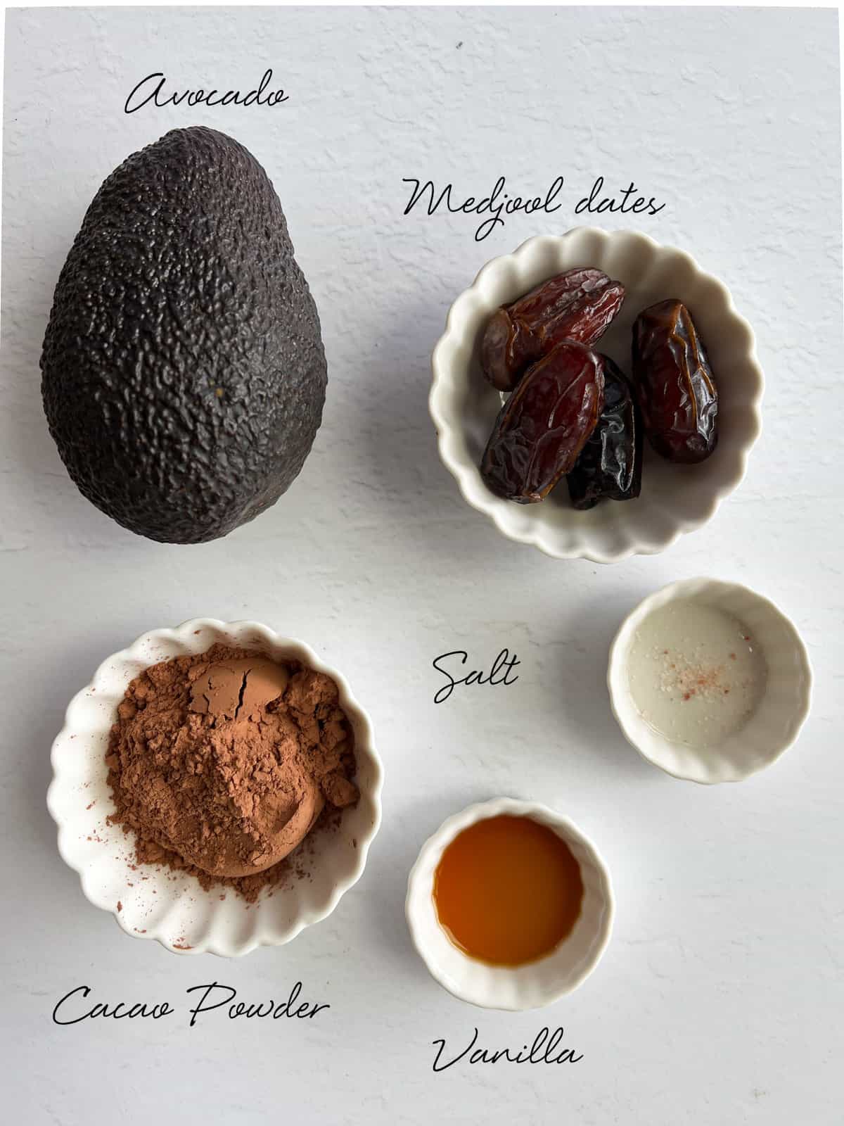 an avocado, dates, cacao powder, salt and vanilla laid out on a white background