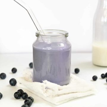 A glass cup with purple milk and blueberries strewn around it