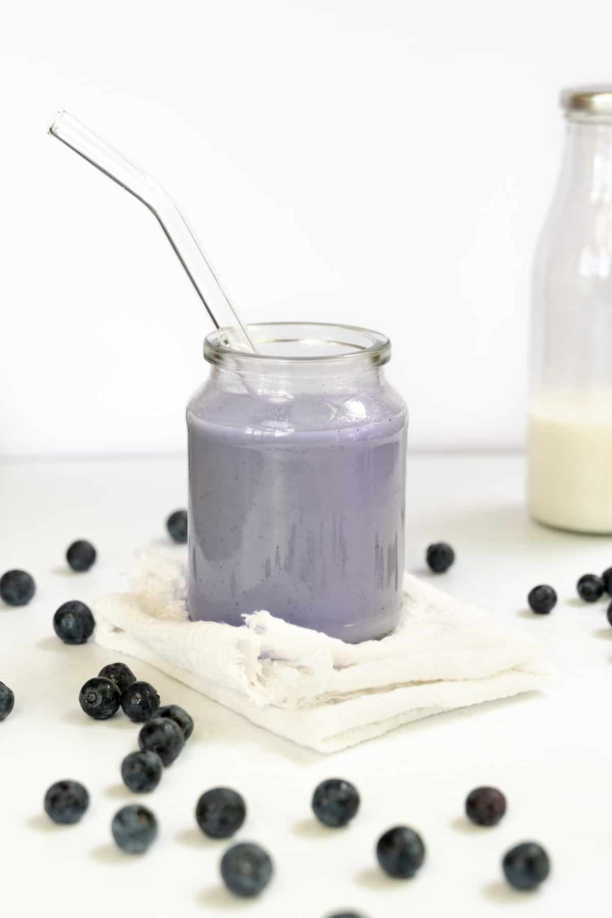 A glass cup with purple milk and blueberries strewn around it