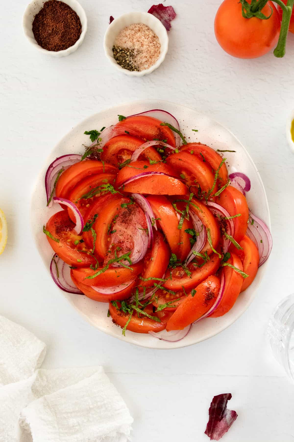 a white plate filled with sliced tomato and onion salad with spices and seasoning on the side