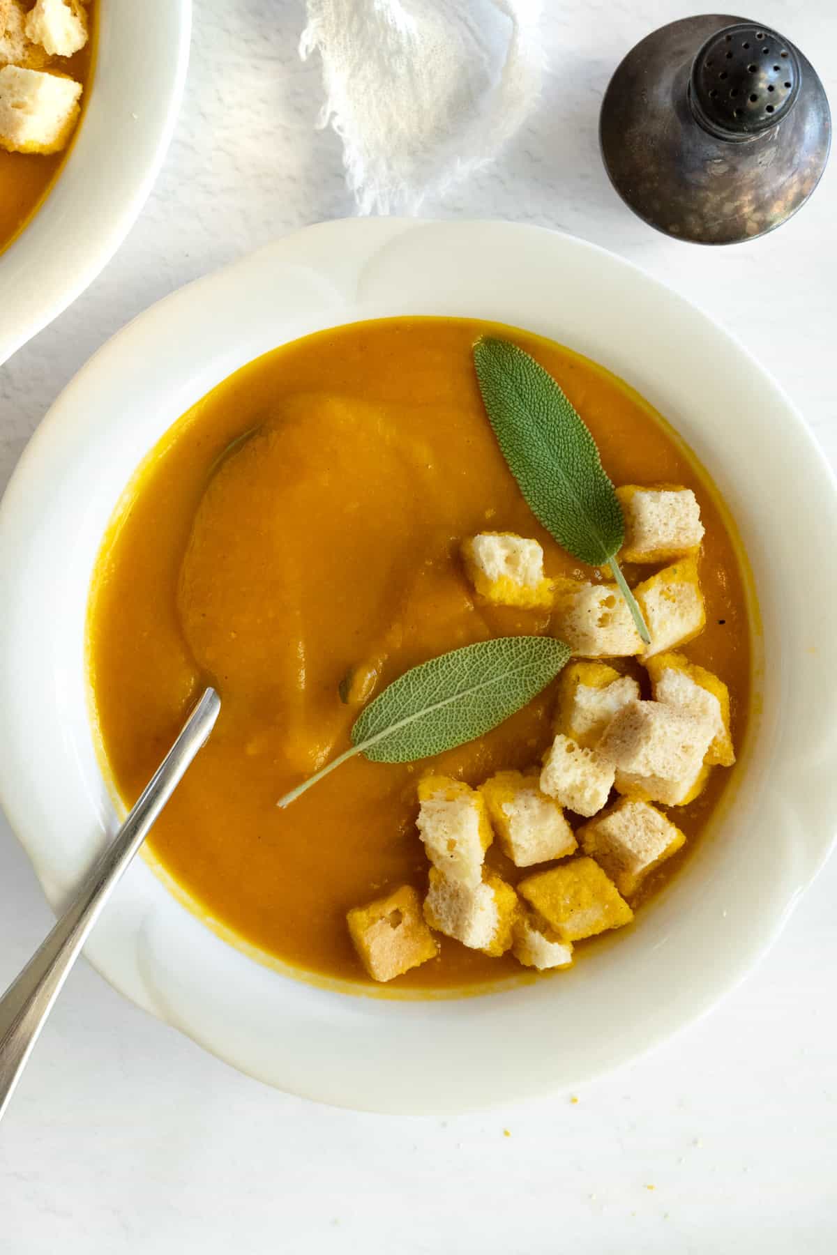 a white bowl with orange soup topped with croutons and a pepper shaker on the side
