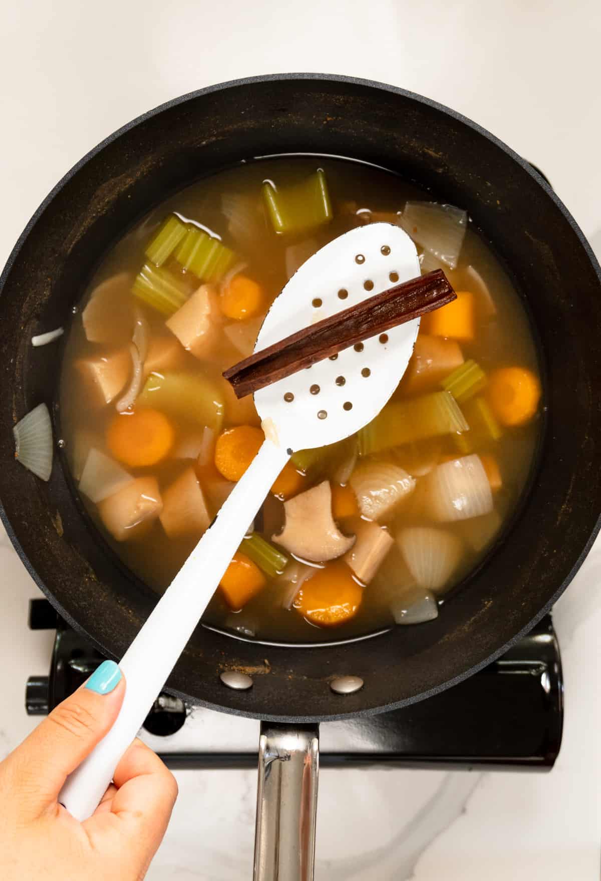 a black pot filled with cooked vegetables and water, with a white slotted spoon holding a cooked cinnamon quill