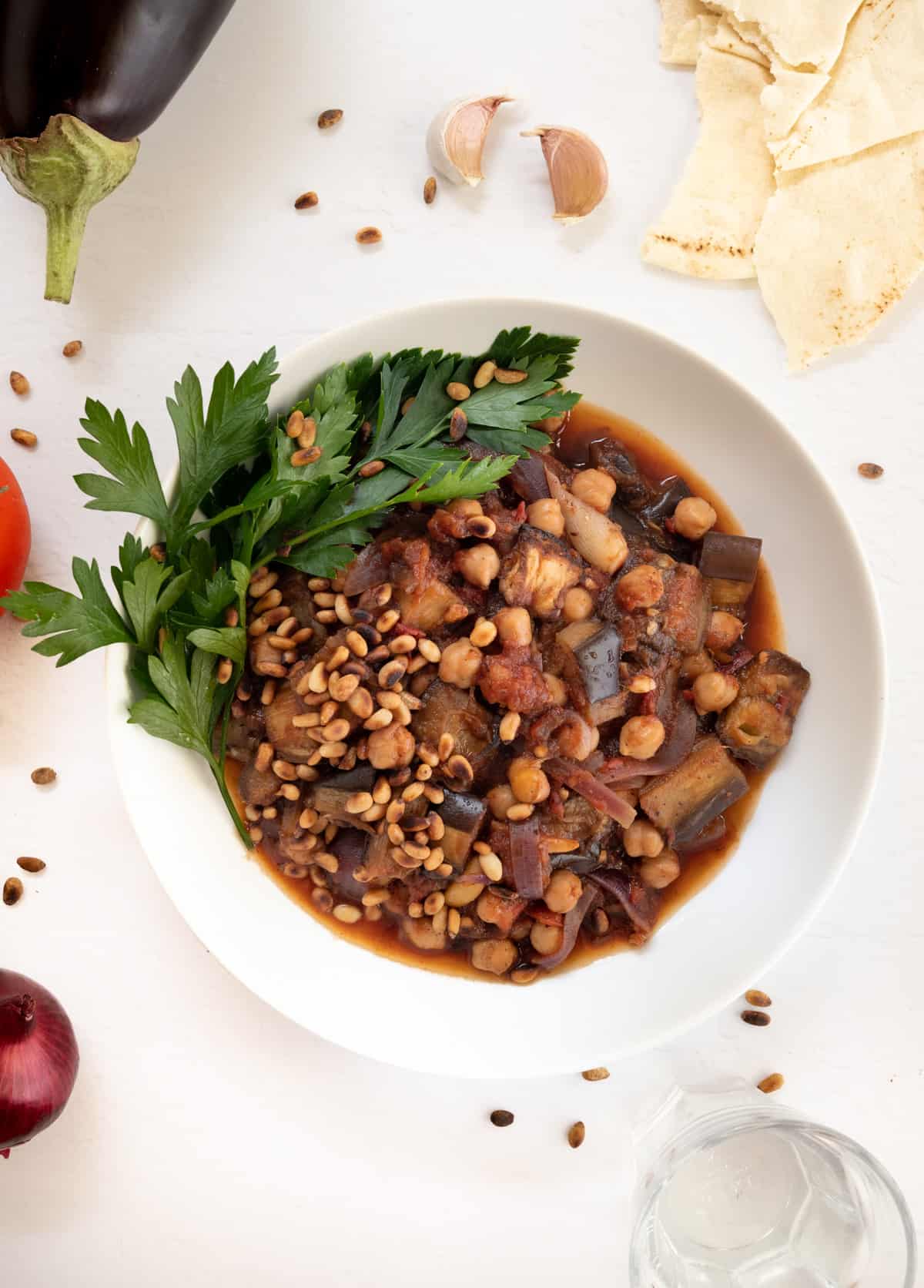 a white plate filled with eggplant, chickpeas and onion cooked in red sauce topped with toasted pine nuts