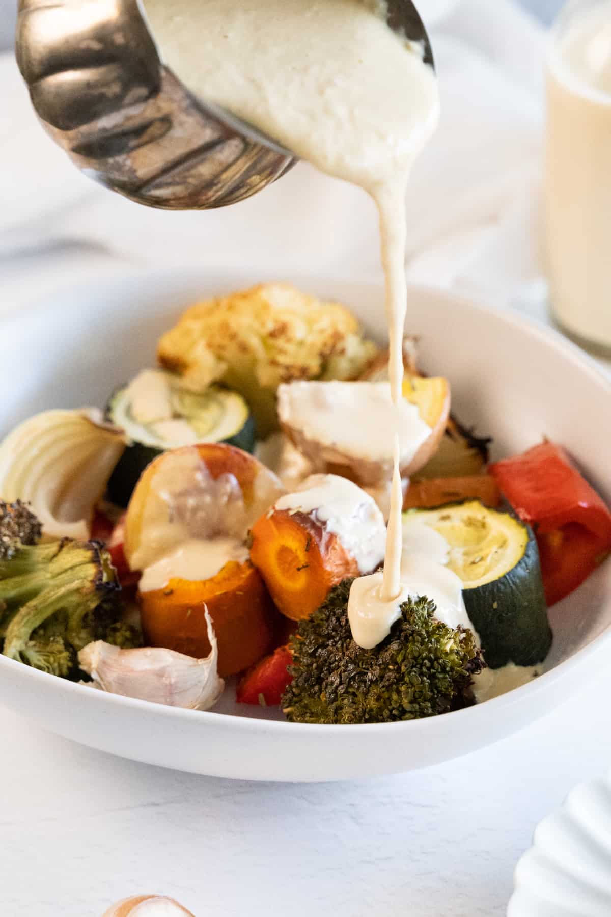 whiteish sauce being poured out of a metal sauce pot over roasted vegetables