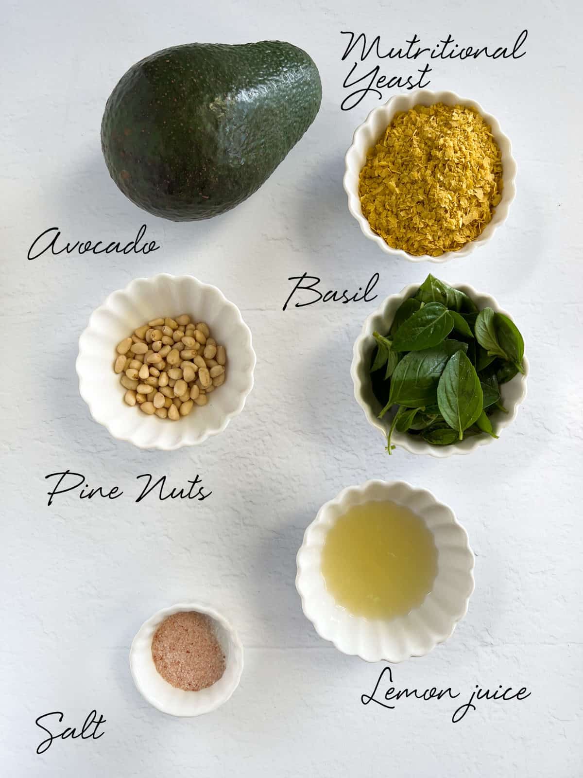 an avocado, nutritional yeast, basil leaves, pine nuts, lemon juice and salt laid out on a white table