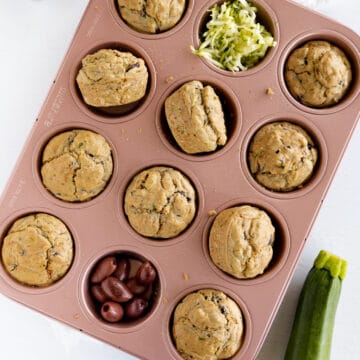 a pink muffin tray filled with 10 muffins, olives and shredded zucchini