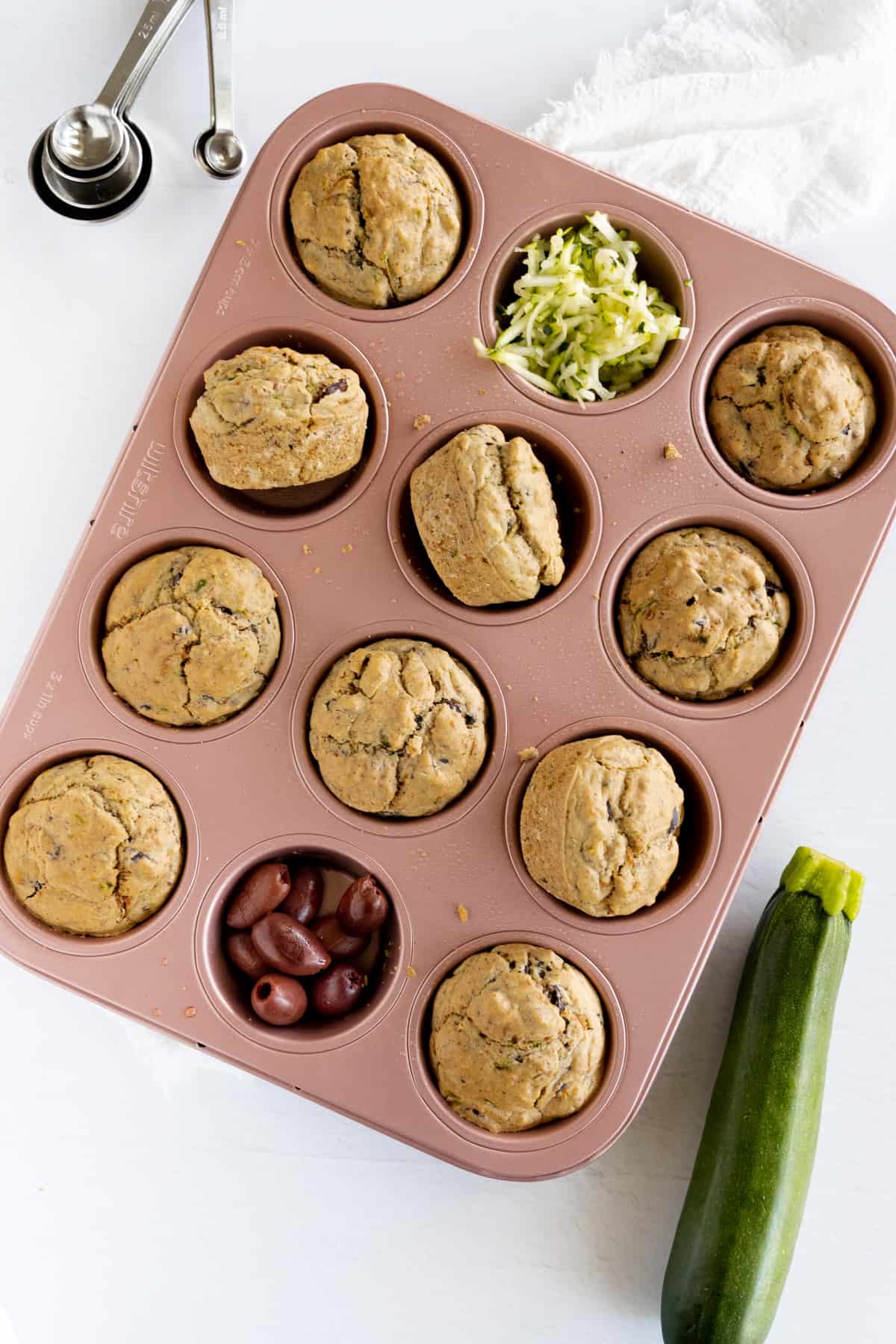 a pink muffin tray filled with 10 muffins, olives and shredded zucchini