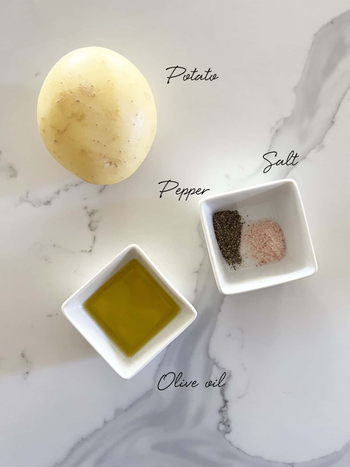 a potato, a bowl of oil and salt and pepper on a white marble bench