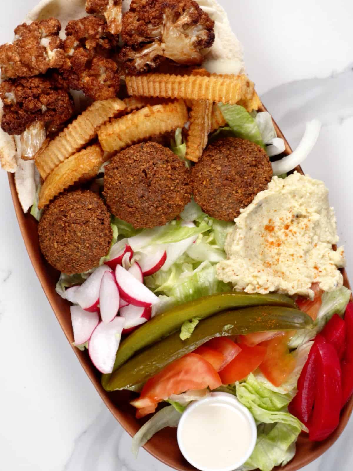 falafel, chopped vegetables, fries, fried cauliflower florets, pink pickles, a dip and sauce and pita bread in a bowl