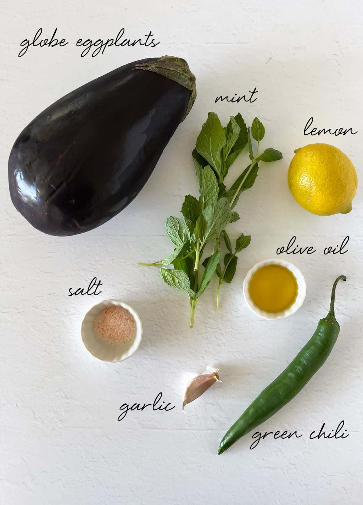a globe eggplant, lemon, mint leaves, salt, green chili, garlic clove and olive oil laid out