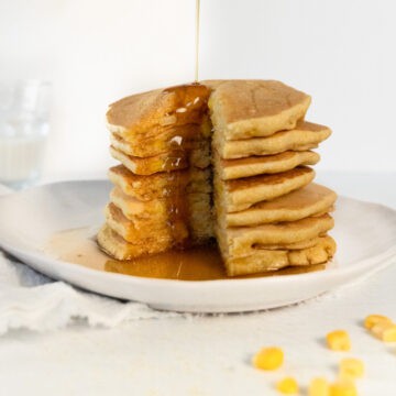 a stack of tall corn pancakes with a slice cut out and maple syrup being poured on top