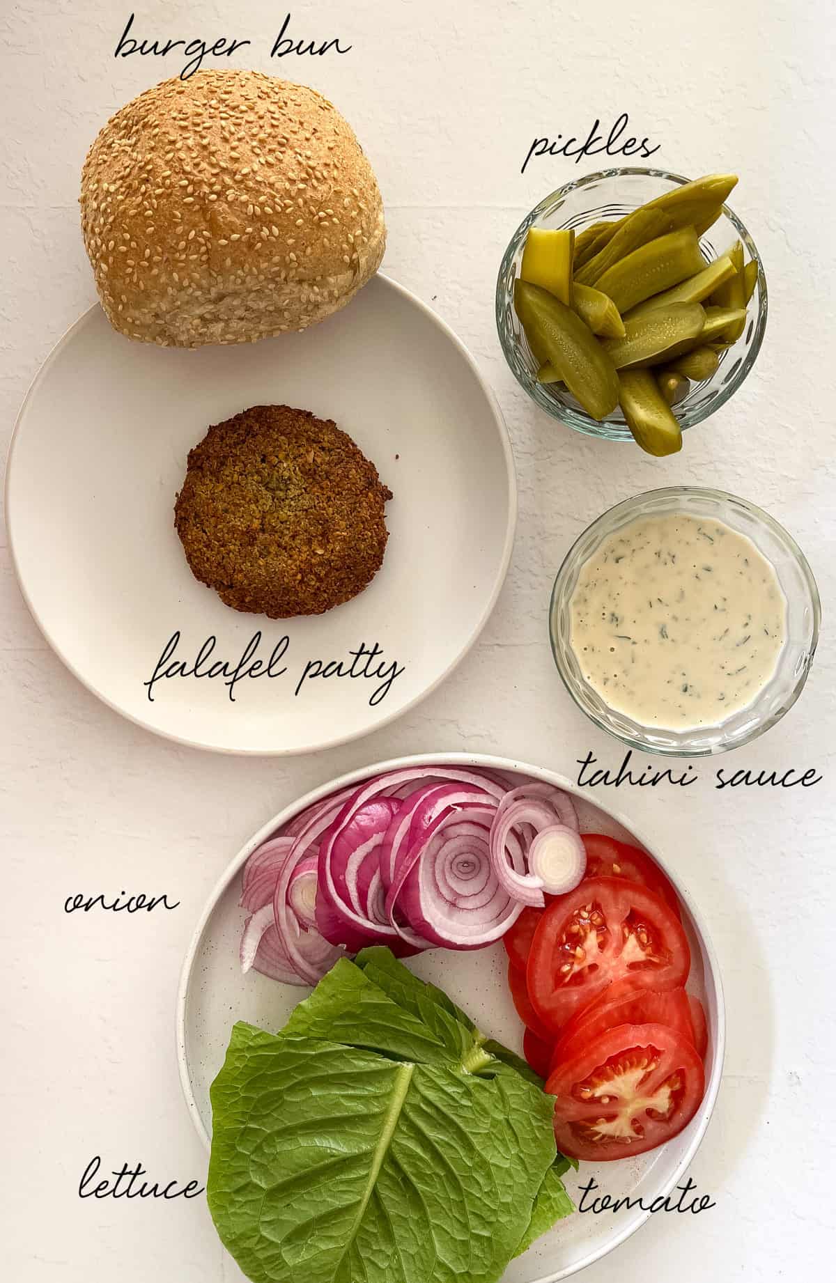 a burger bun, falafel patty, lettuce, tomato, onion, tahini dressing, pickled cucumber laid out