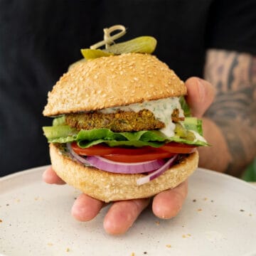 a male hand holding a vegan falafel burger with tahini dripping from it