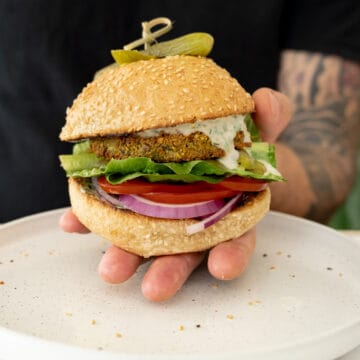 a male hand holding a vegan falafel burger with tahini dripping from it