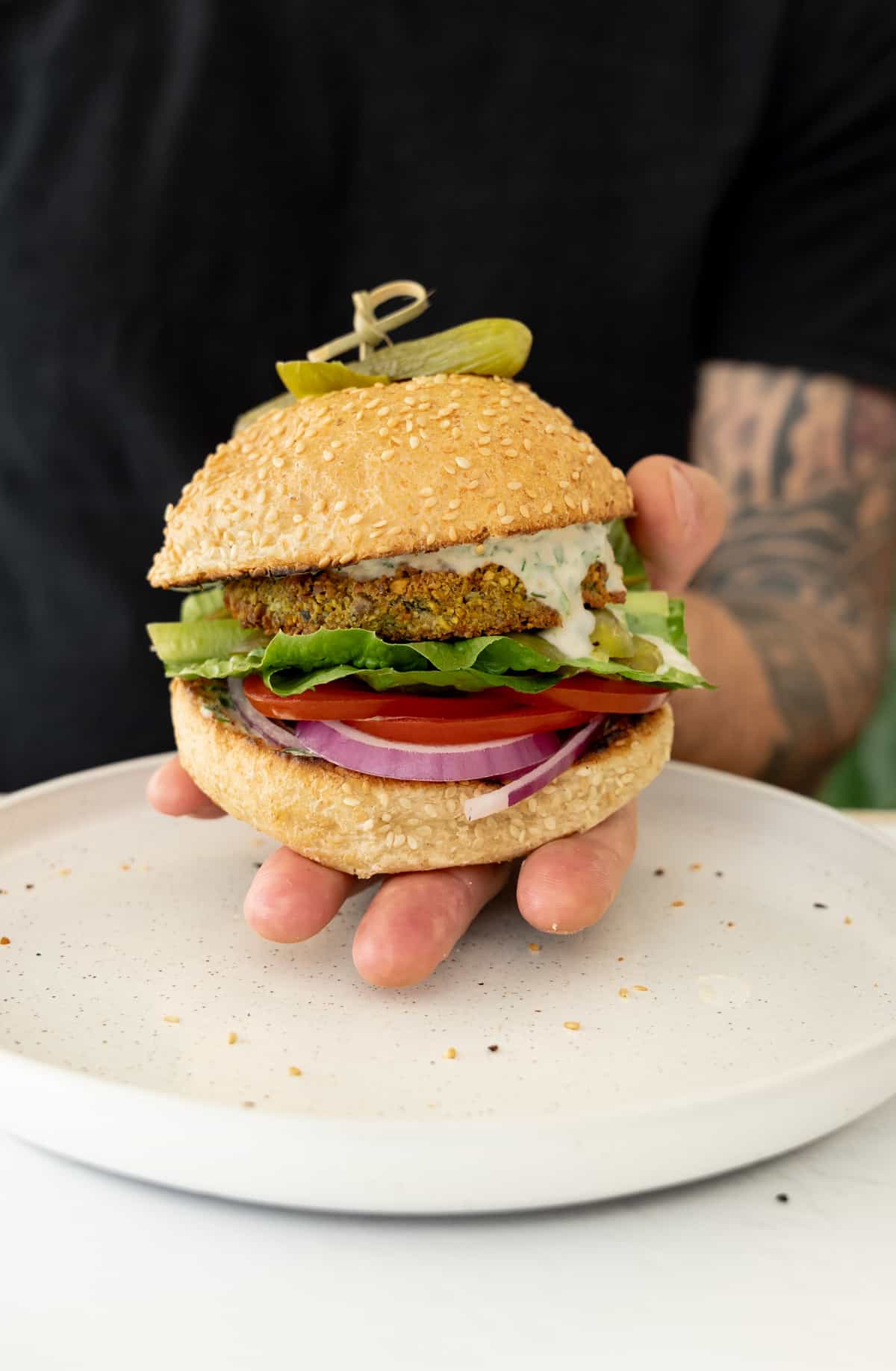 a male hand holding a vegan falafel burger with tahini dripping from it
