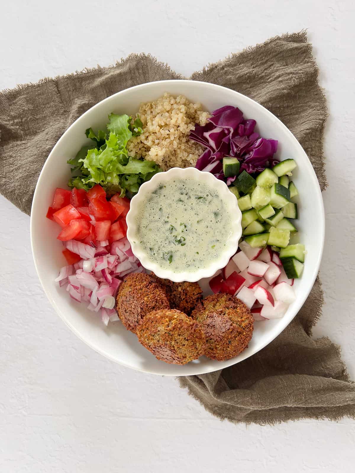 falafel balls, diced veggies and chopped lettuce in a plate with a bowl of lemon tahini dressing in the centre