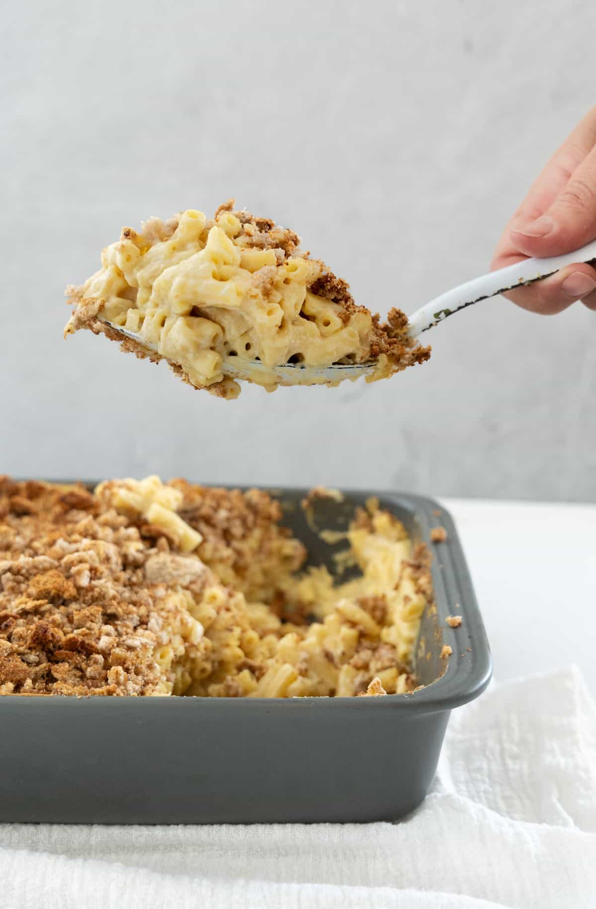 a hand lifting a large spoon with vegan baked mac and cheese in it on top of a casserole dish filled with it